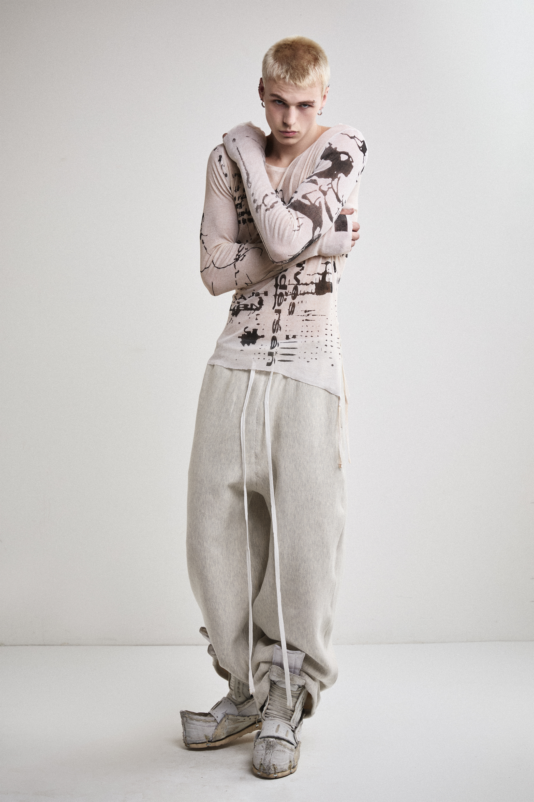 Front view of Tyler Andersen wearing a sheer long-sleeve cream AOA top with black text and graphics designed to look like tattoos.