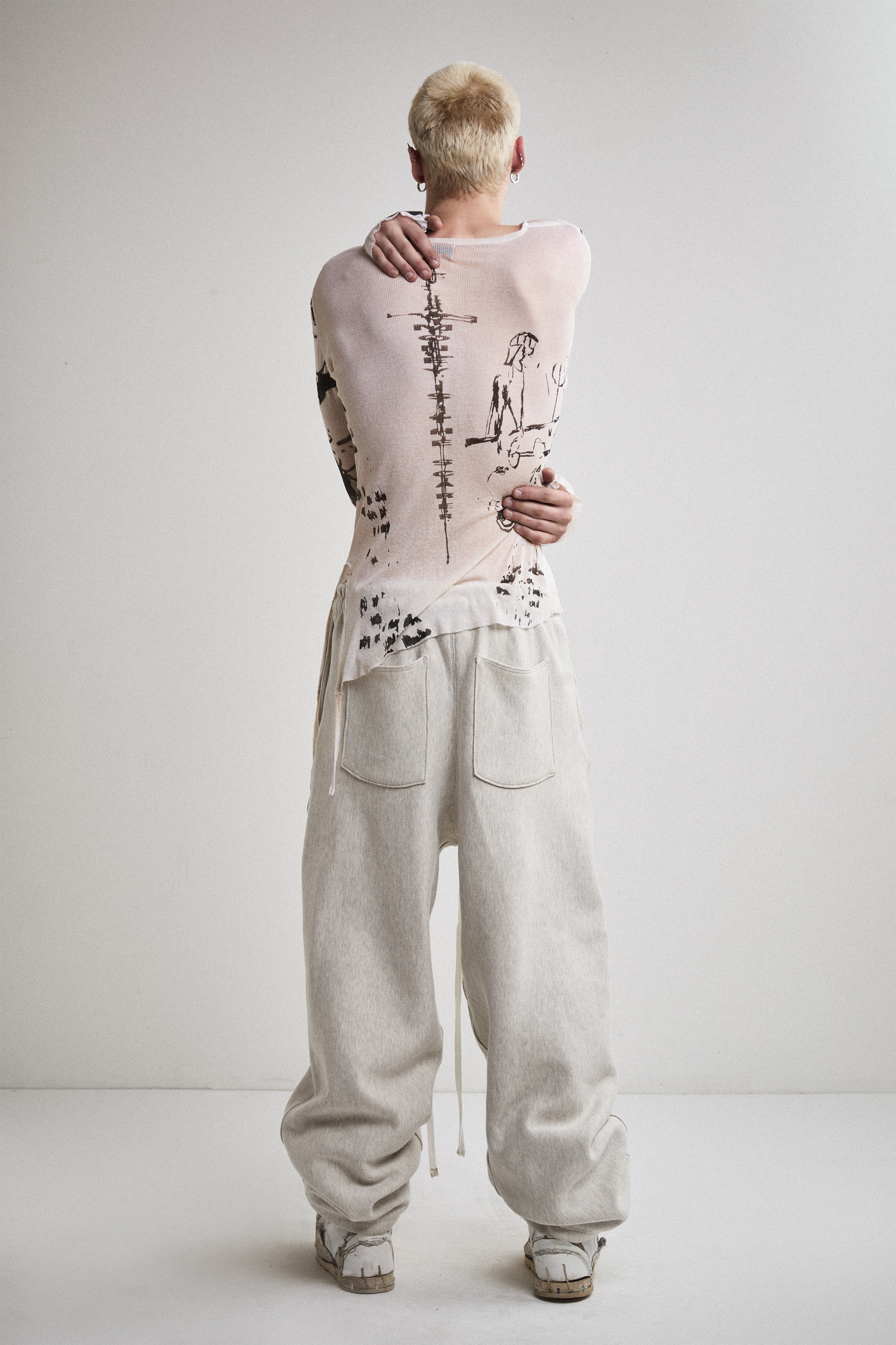 Back view of Tyler Andersen with hands showing wearing a sheer long-sleeve cream AOA top with black text and graphics designed to look like tattoos.