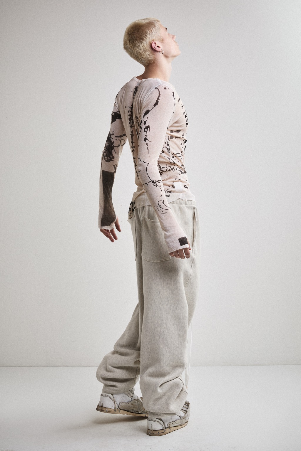 Side view of Tyler Andersen wearing a sheer long-sleeve cream AOA top with black text and graphics designed to look like tattoos.