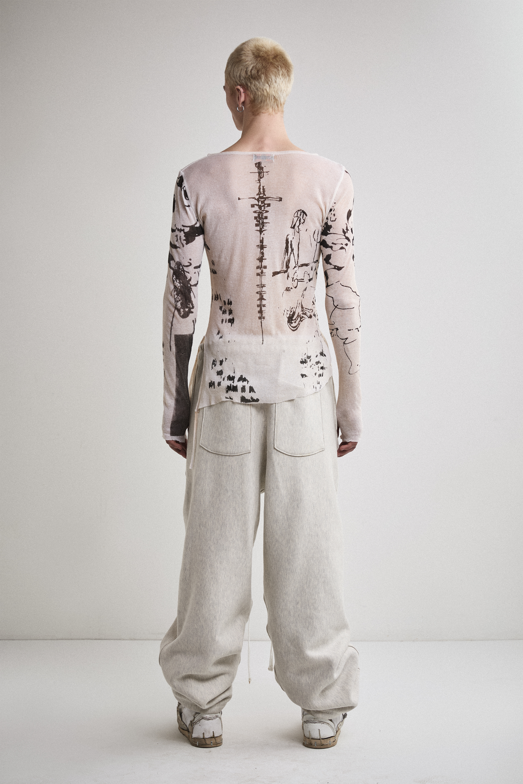 Back view of Tyler Andersen wearing a sheer long-sleeve AOA cream top with black text and graphics designed to look like tattoos.