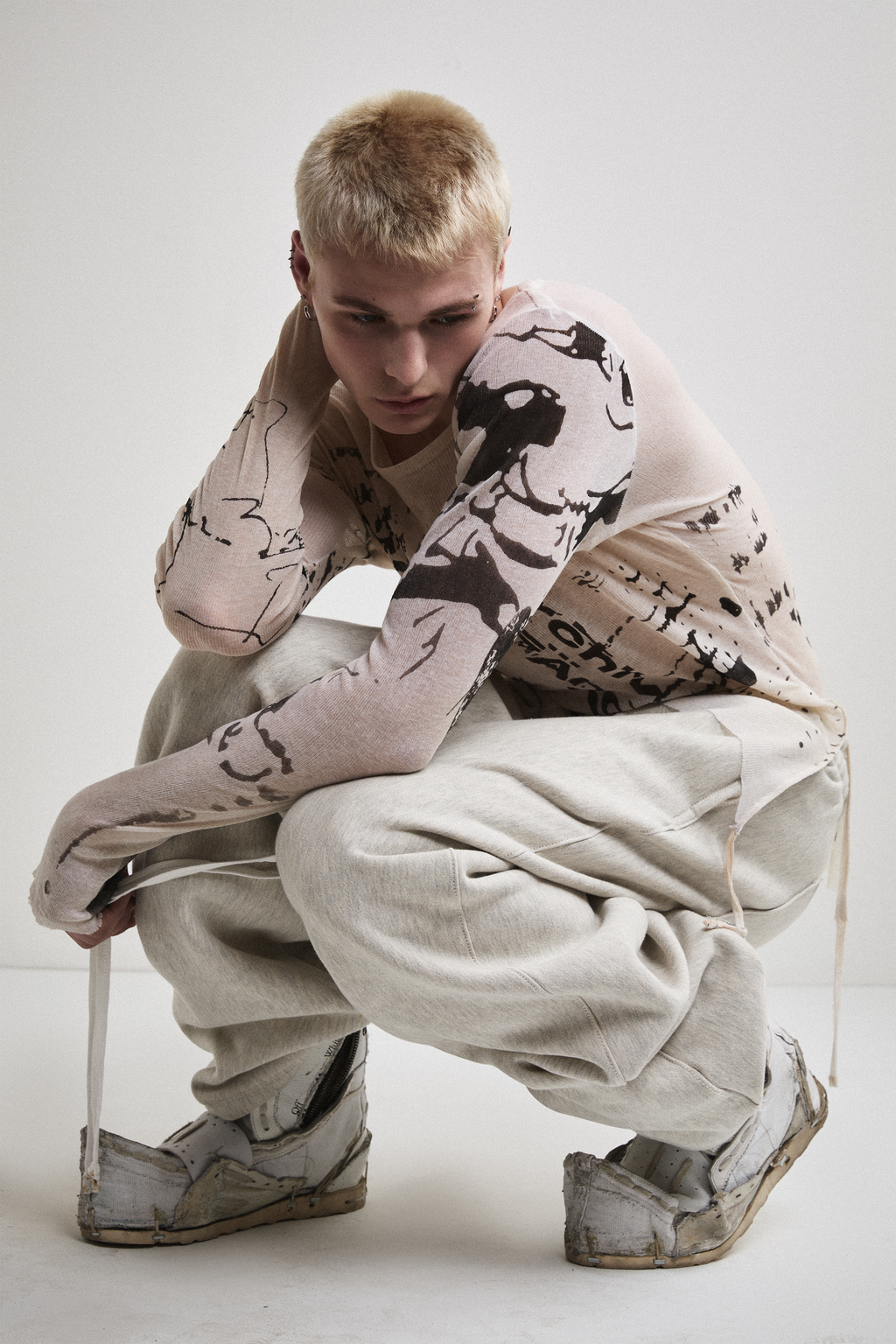 Front view of Tyler Andersen crouched wearing a sheer long-sleeve cream AOA top with black text and graphics designed to look like tattoos.