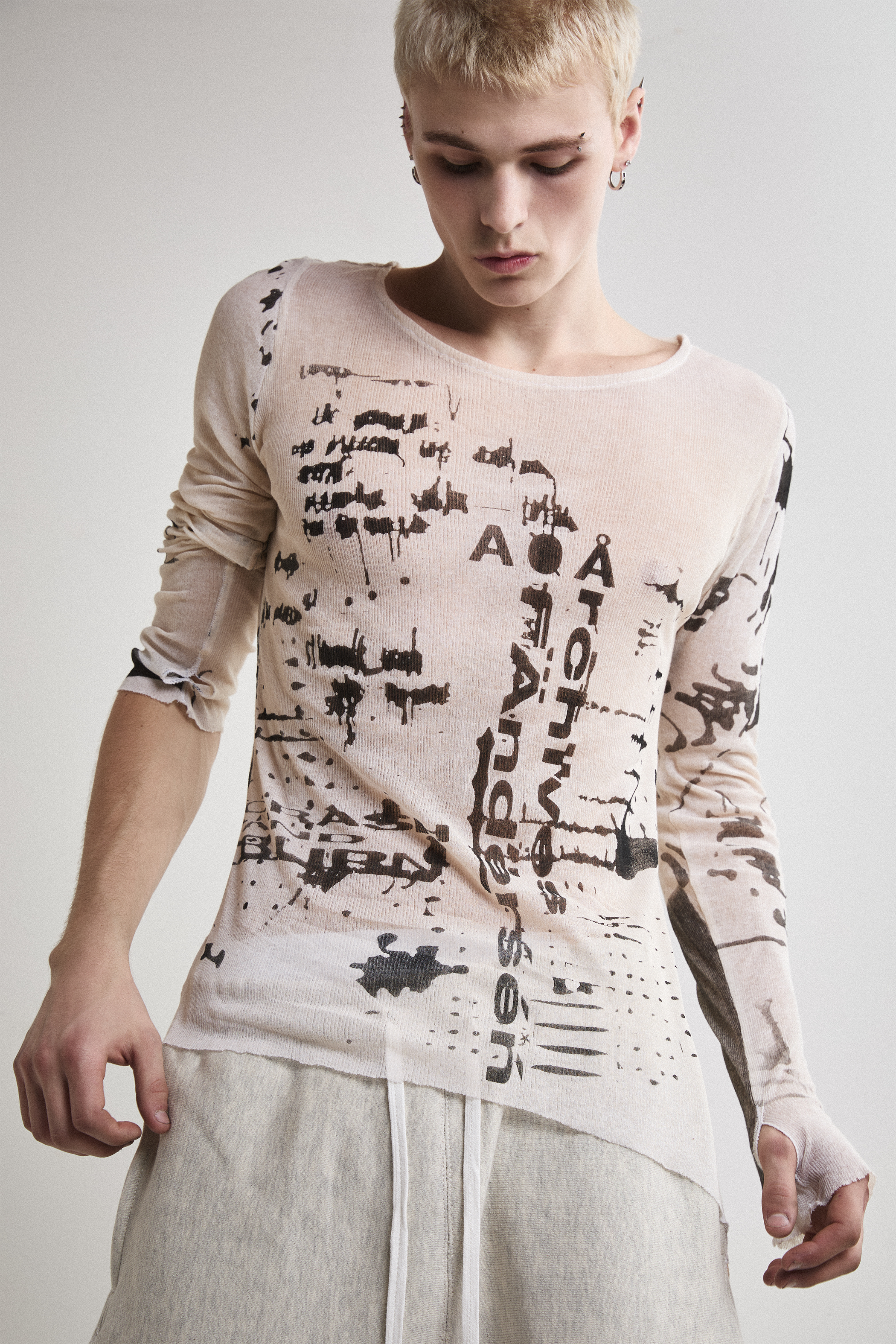 Front close-up view of Tyler Andersen wearing a sheer long-sleeve cream AOA top with black text and graphics designed to look like tattoos.