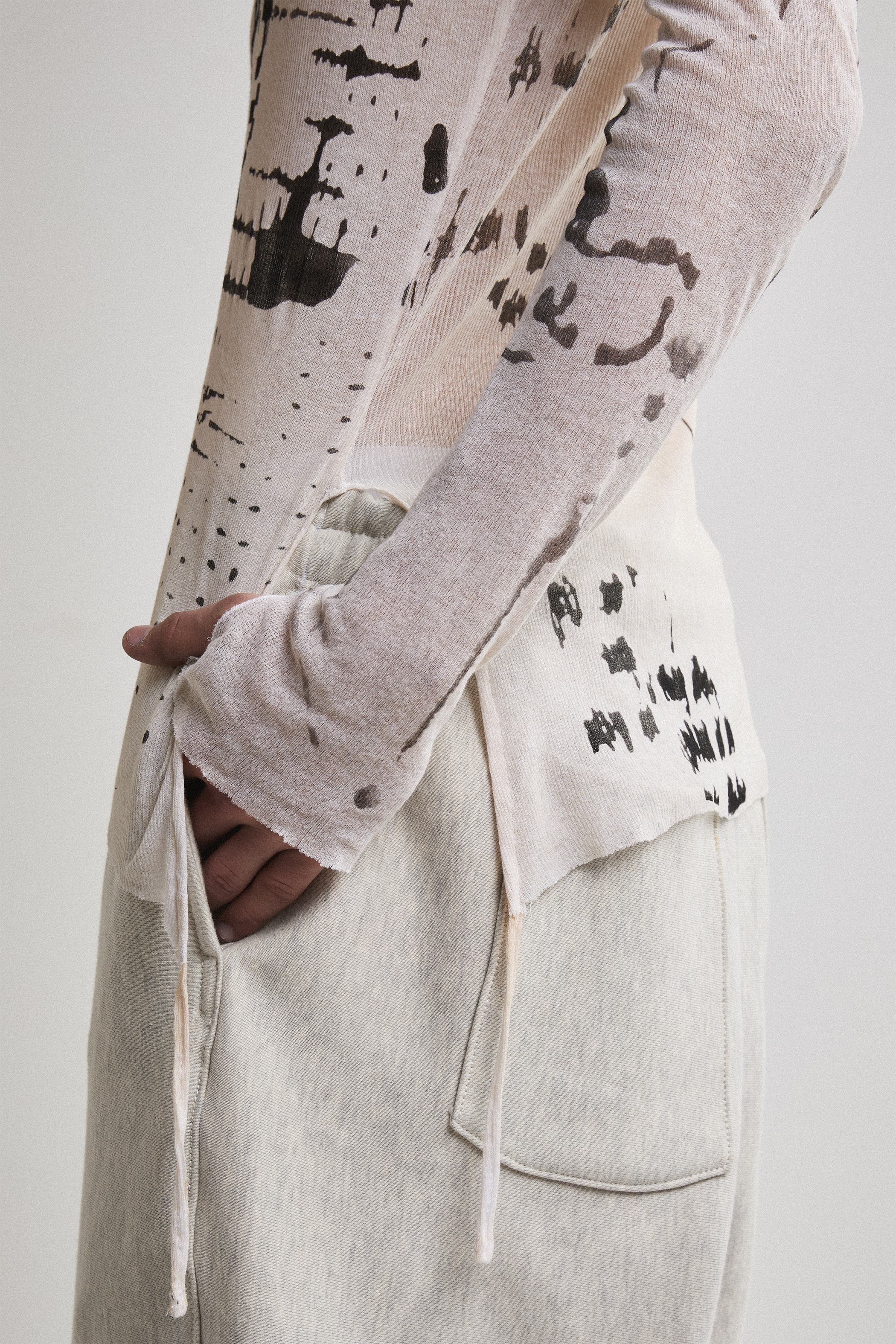 Close-up side view of Tyler Andersen wearing a sheer long-sleeve cream AOA top with black text and graphics designed to look like tattoos with hands in pocket.