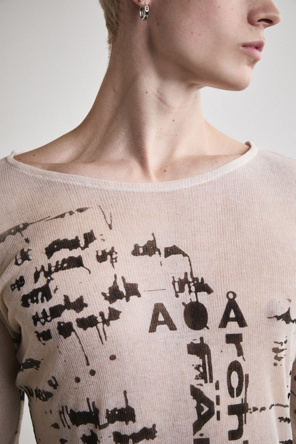 Front view of Tyler Andersen looking sideways wearing a sheer long-sleeve cream AOA top with black text and graphics designed to look like tattoos.