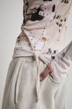 Close-up side view of Tyler Andersen wearing a sheer long-sleeve cream AOA top with black text and graphics designed to look like tattoos showing thumb holes and tassels.