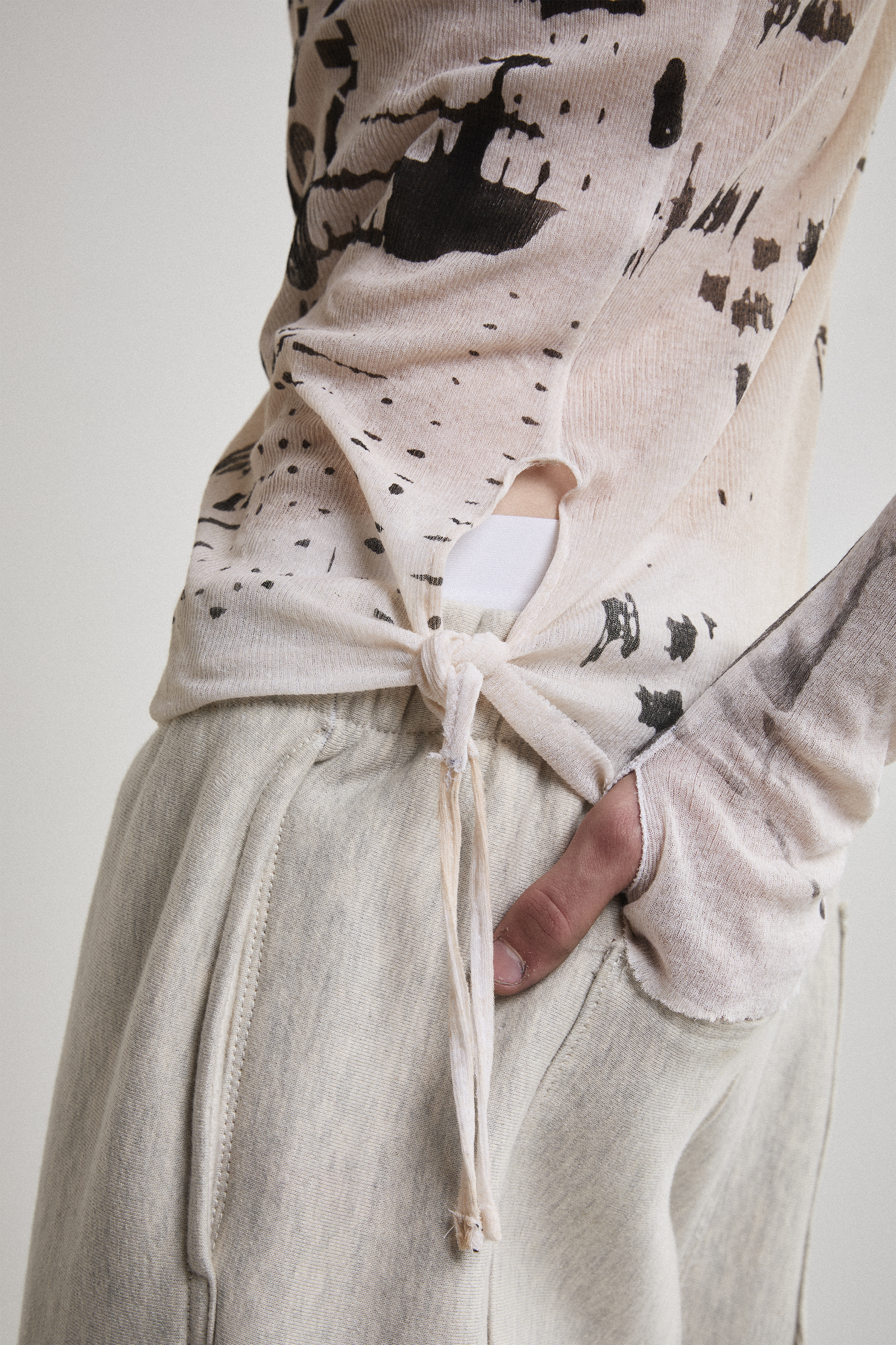 Close-up side view of Tyler Andersen wearing a sheer long-sleeve cream AOA top with black text and graphics designed to look like tattoos showing thumb holes and tassels.