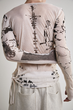 Close-up back view with arm crossed of Tyler Andersen wearing a sheer long-sleeve cream AOA top with black text and graphics designed to look like tattoos.