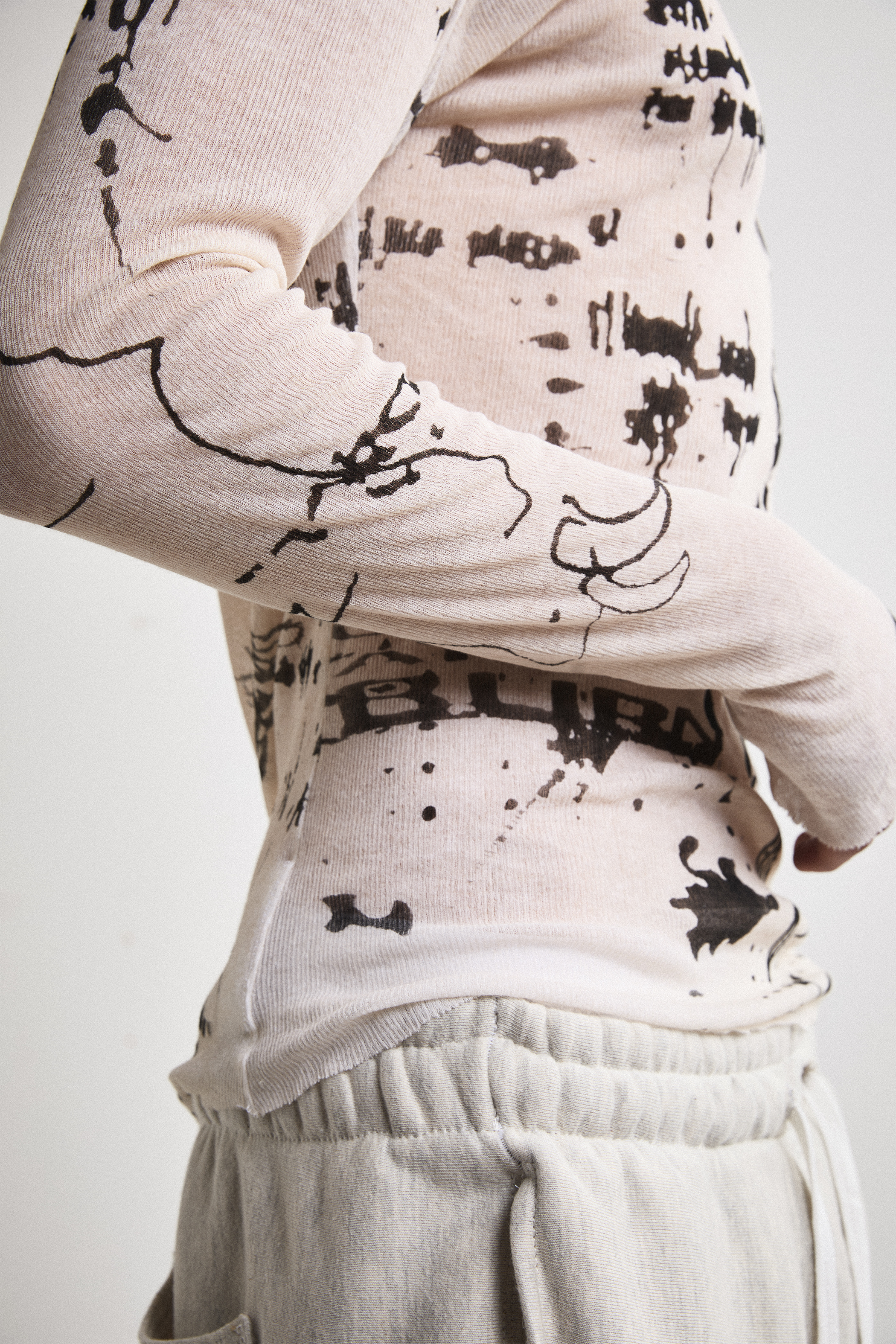 Close-up right side view of Tyler Andersen wearing a sheer long-sleeve cream AOA top with black text and graphics designed to look like tattoos.