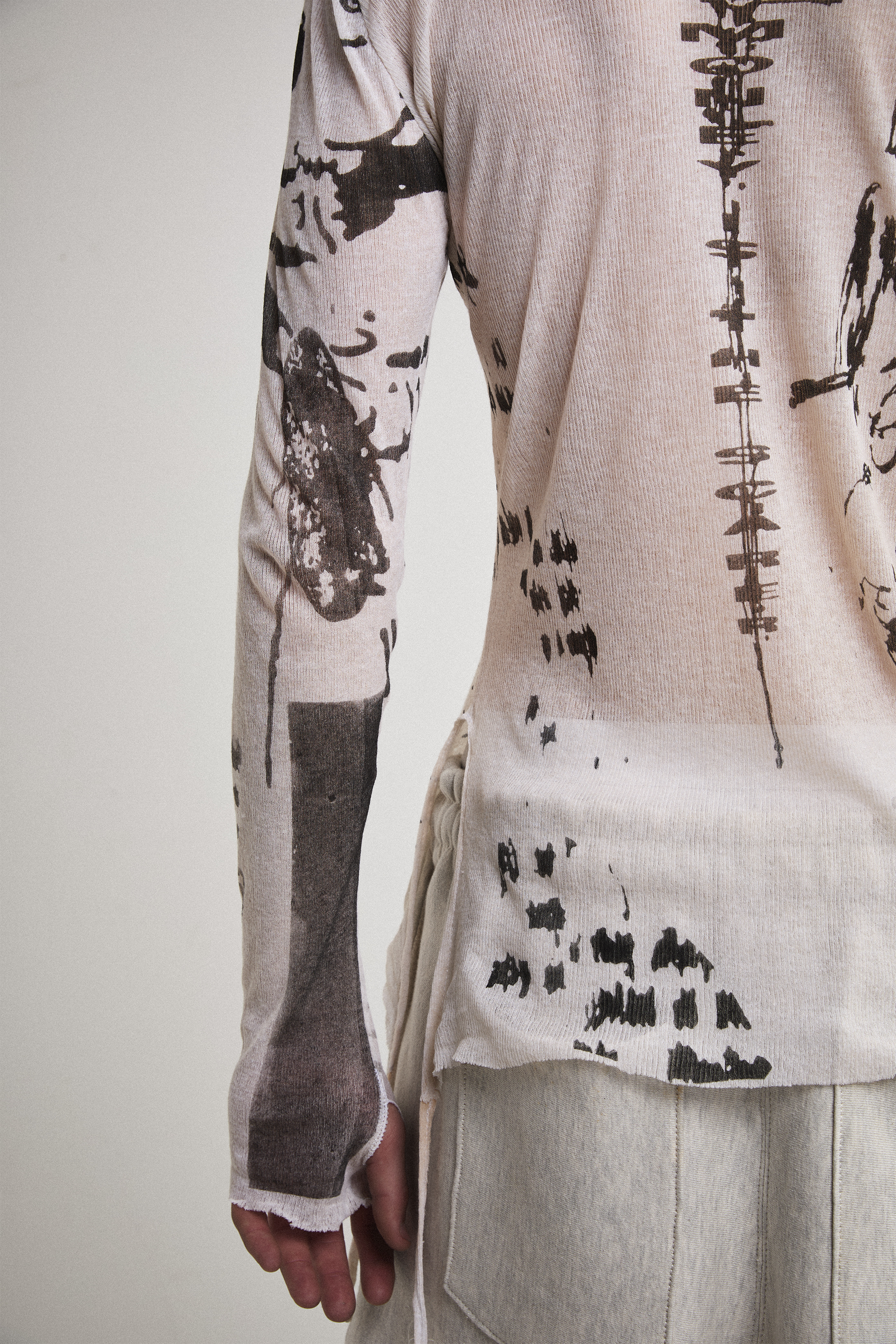 Close-up left side view of Tyler Andersen wearing a sheer long-sleeve cream AOA top with black text and graphics designed to look like tattoos.