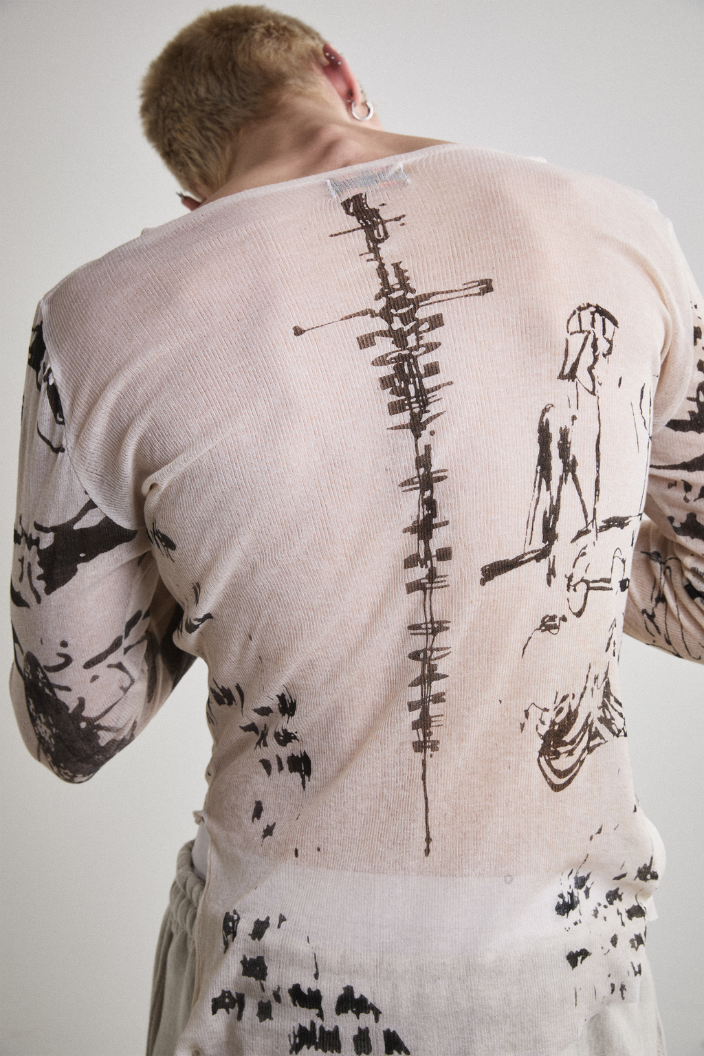 Close-up back view of Tyler Andersen wearing a sheer long-sleeve cream AOA top with black text and graphics designed to look like tattoos.