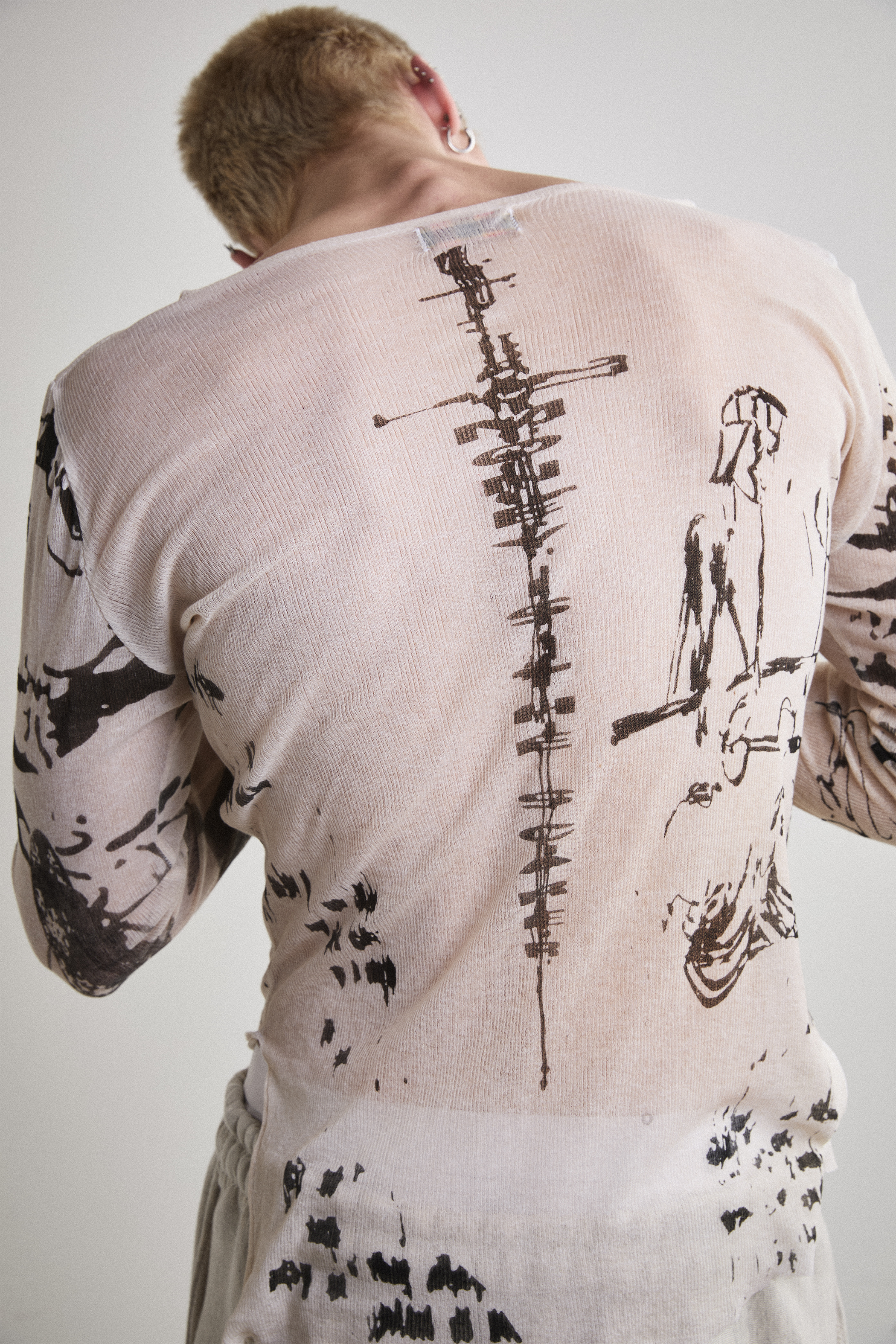 Close-up back view of Tyler Andersen wearing a sheer long-sleeve cream AOA top with black text and graphics designed to look like tattoos.